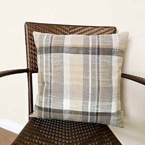 May include: A square throw pillow featuring a plaid pattern in shades of beige, tan, white, gray, and charcoal. The fabric has a visible woven texture with intersecting horizontal and vertical lines forming a classic grid design across the surface.