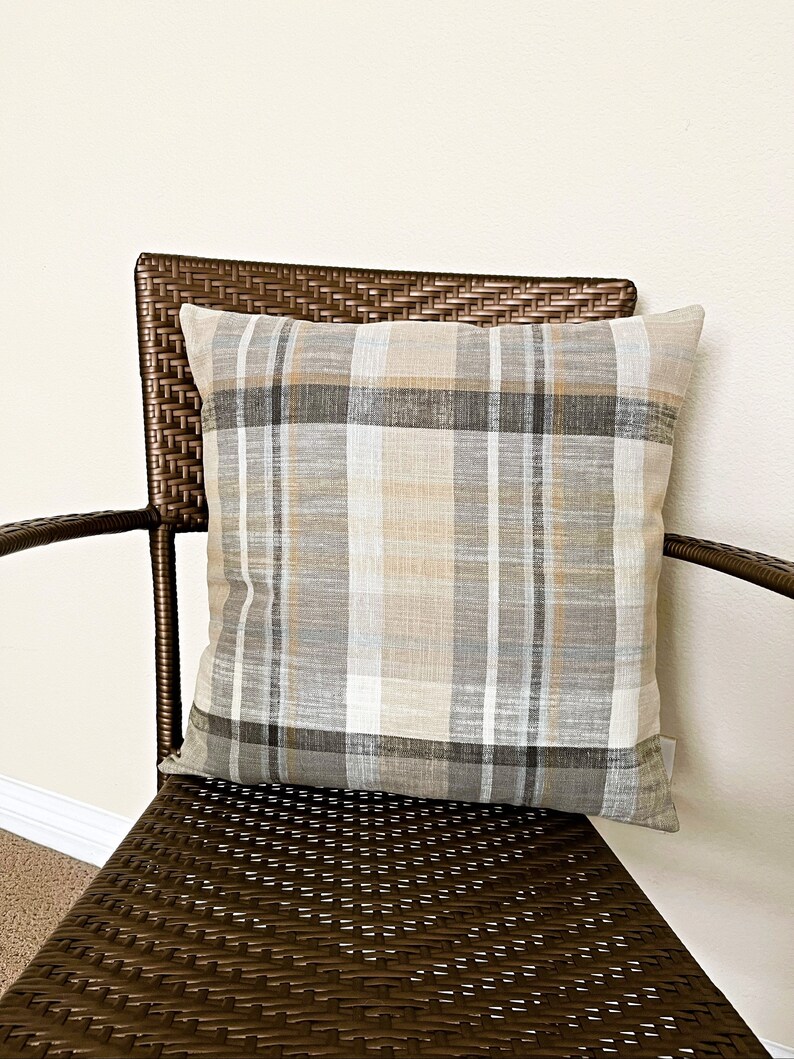 May include: A square throw pillow featuring a plaid pattern in shades of beige, tan, white, gray, and charcoal. The fabric has a visible woven texture with intersecting horizontal and vertical lines forming a classic grid design across the surface.