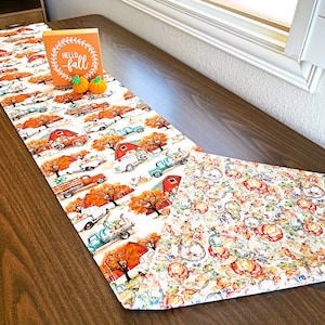 Peut inclure: Un chemin de table blanc sur le thème de l'automne. Un côté présente un motif de granges rouges, de camions vintage et d'arbres d'automne. L'autre côté présente un motif de citrouilles, de courges et de feuilles. Le chemin de table est sur une table en bois avec une pancarte qui dit "Hello Fall".