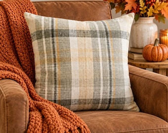 May include: A square throw pillow featuring a plaid pattern in shades of sage green, cream, and mustard yellow. The fabric has a visible woven texture, and the pillow is displayed against a brown leather chair with a chunky orange knit throw.