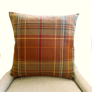 Pode incluir: Uma almofada quadrada com uma cor de base castanha e um padr&atilde;o de xadrez multicolorido. O design consiste em linhas horizontais e verticais que se cruzam em tons de vermelho, amarelo, azul e verde, criando uma grelha de estilo tartan.