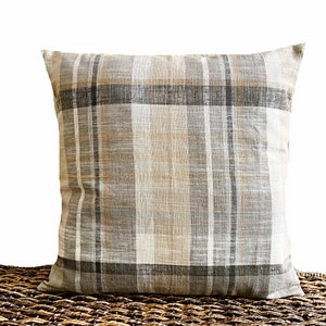 May include: A square throw pillow featuring a woven plaid pattern in neutral shades of beige, tan, gray, and white. The fabric has a visible textured weave, and the design consists of intersecting vertical and horizontal stripes of varying widths.