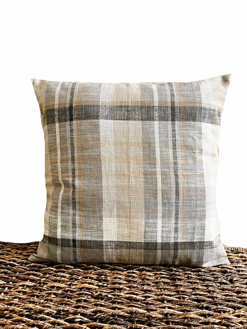 May include: A square throw pillow featuring a woven plaid pattern in neutral shades of beige, tan, gray, and white. The fabric has a visible textured weave, and the design consists of intersecting vertical and horizontal stripes of varying widths.