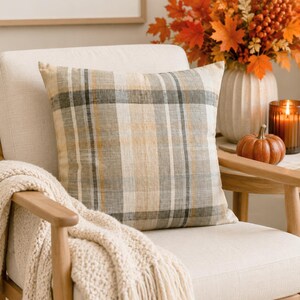 May include: A square throw pillow featuring a plaid pattern in shades of beige, gray, and muted orange. The fabric has a visible woven texture, and the pillow is positioned on a light-colored upholstered chair with a cream-colored knit throw blanket.