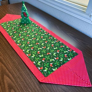 May include: A green and red Christmas table runner with a pattern of stockings, candy canes, and holly leaves. The runner has a red border with white polka dots.