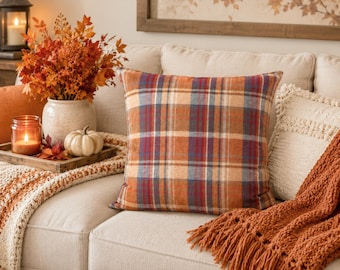 May include: A square throw pillow featuring a plaid pattern in shades of burnt orange, deep red, navy blue, and cream. The fabric has a soft, textured appearance, and the pillow is displayed in a centered position, showcasing the intersecting horizontal and vertical lines of the classic tartan design.