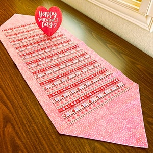 May include: A pink and red table runner with a heart pattern and the text "Happy Valentine's Day" on a red heart-shaped sign.