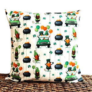 May include: A white pillow with a green, orange, and gold pattern of leprechauns, cars, pots of gold, and shamrocks. The pillow is on a woven brown surface.