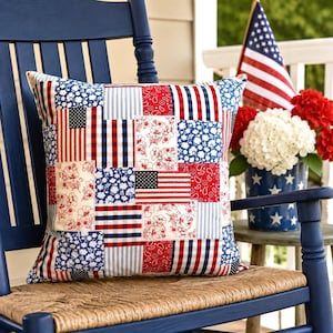 May include: A square pillow with a patchwork design featuring red, white, and blue patterns, including American flags, floral prints, and stripes. The pillow rests on a blue rocking chair with a woven seat.