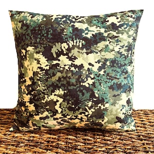 May include: Square decorative pillow with a camouflage pattern in shades of green, blue, and beige. The pillow is placed on a woven surface, creating a contrast in textures. The pillow's design resembles a forest or woodland scene.