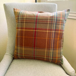 Pode incluir: Uma almofada quadrada com um padr&atilde;o xadrez multicolorido. O tecido apresenta uma base em tons de castanho quente, cruzada por linhas finas verticais e horizontais em tons de vermelho, amarelo, azul e verde claro, criando uma grelha.