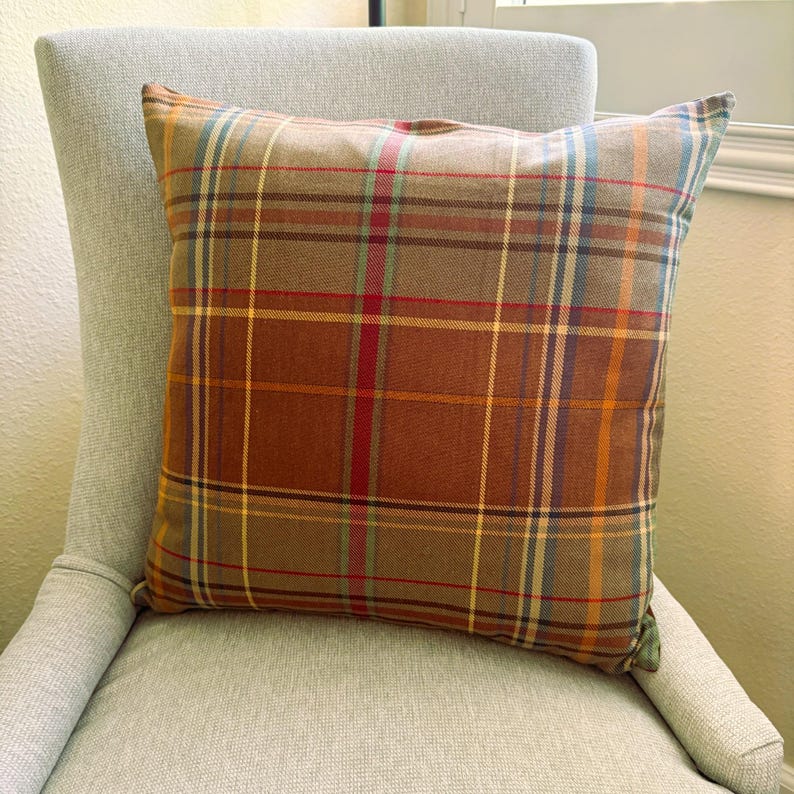 Pode incluir: Uma almofada quadrada com um padr&atilde;o xadrez multicolorido. O tecido apresenta uma base em tons de castanho quente, cruzada por linhas finas verticais e horizontais em tons de vermelho, amarelo, azul e verde claro, criando uma grelha.
