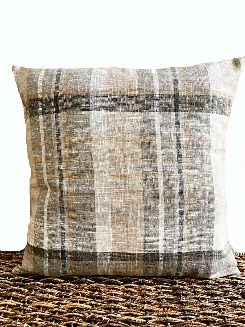 May include: A square throw pillow featuring a woven plaid pattern in neutral shades of beige, light gray, dark charcoal, and soft white. The fabric has a visible textured weave, and the design consists of intersecting vertical and horizontal stripes.