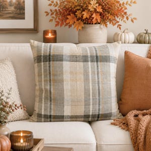 May include: A square throw pillow featuring a woven plaid pattern in neutral shades of beige, cream, gray, and muted brown. The fabric has a visible linen-like texture with intersecting vertical and horizontal lines forming a classic grid design.