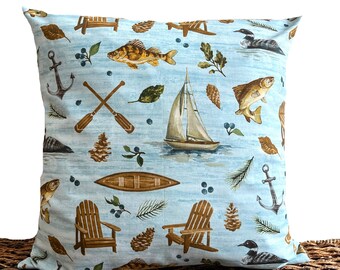 Pode incluir: Uma almofada azul com uma variedade de ilustrações com tema natural, incluindo peixes, barcos, canoas e pinhas. A almofada tem um fundo azul claro com um efeito de aquarela sutil.