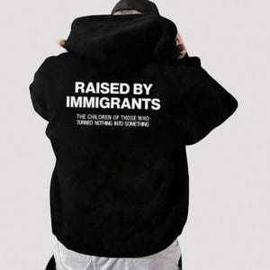 Pode incluir: Moletom preto com a frase branca "RAISED BY IMMIGRANTS". Abaixo, a frase "THE CHILDREN OF THOSE WHO TURNED NOTHING INTO SOMETHING". O moletom é feito de um material macio.