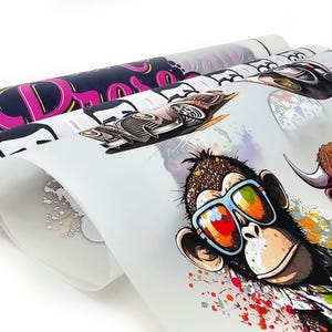 May include: Rolled sheets of printed designs featuring a monkey wearing sunglasses, a vintage car, and other graphics. The monkey has a colorful splatter design around it. The designs are printed on a white background.