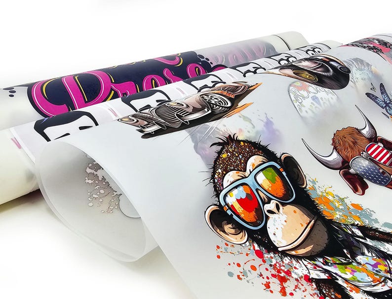 May include: Rolled sheets of printed designs featuring a monkey wearing sunglasses, a vintage car, and other graphics. The monkey has a colorful splatter design around it. The designs are printed on a white background.