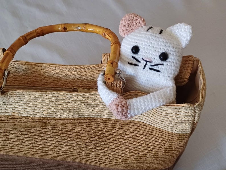 May include: A white crocheted cat toy with pink ears peeking out of a brown and beige woven tote bag with a bamboo handle.