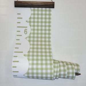 May include: A wall-hanging growth chart featuring a sage green and white gingham pattern. The chart has a scalloped white edge with black measurement markings and the number 6 printed on it. It is suspended from a dark brown wooden frame with twine.