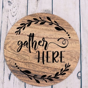 May include: Round wooden sign with the text "gather here" in black script lettering. The sign is decorated with a black floral wreath.