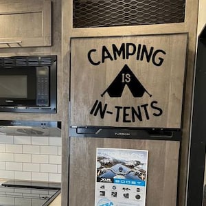 May include: A refrigerator with a wooden door featuring the text "CAMPING IS IN-TENTS" in black. A microwave and range hood are visible to the left. A flyer is attached to the refrigerator door.