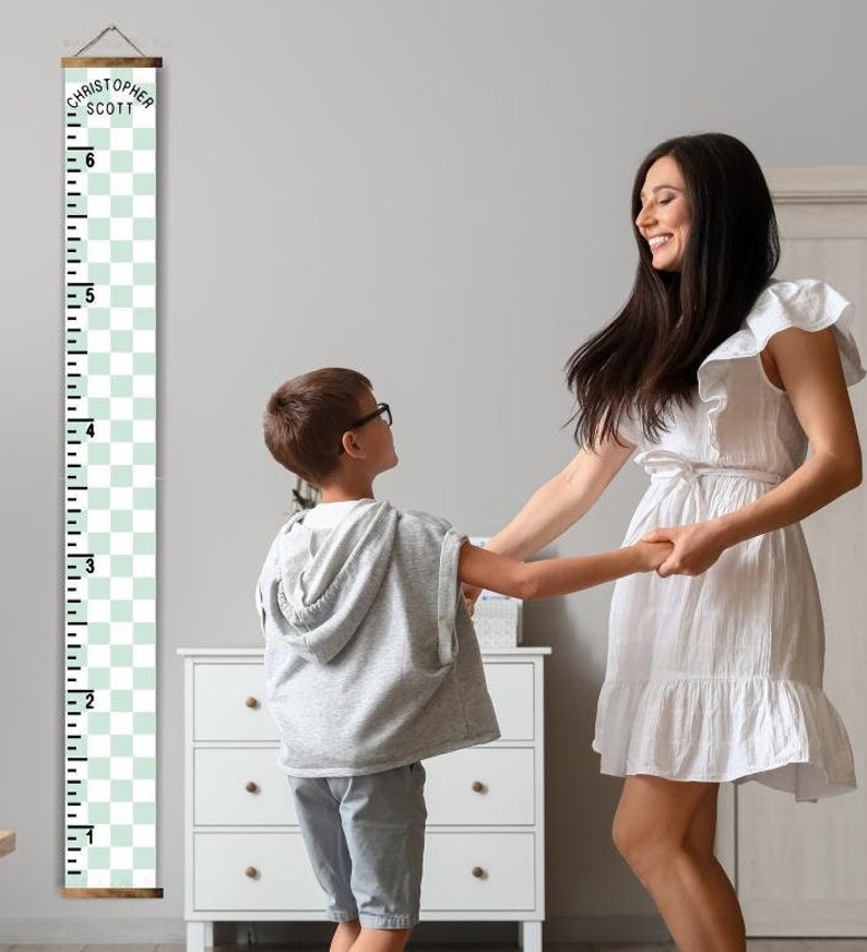 May include: A green and white checkered growth chart with a wooden frame and the name "Christopher Scott" printed at the top. A child and an adult are standing next to the chart, holding hands and smiling.