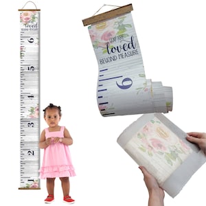 May include: A white wooden growth chart with floral accents and the text "You are loved beyond measure". The chart is rolled up and has a wooden dowel at the top. A child is standing next to the chart.