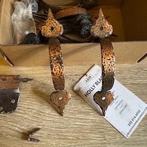 以下が含まれることがあります： ハート型のデザインが施された、装飾的な銅製のドアヒンジが2つ。ヒンジは、白いラベルが貼られた箱に入っています。ラベルには「Holly Black」と連絡先情報が記載されています。