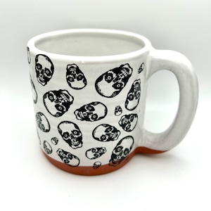 Könnte beinhalten: Weiße Keramiktasse mit einem schwarzen Totenkopf-Muster. Die Tasse hat einen braunen Rand und einen Henkel.