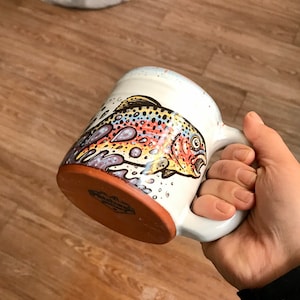 May include: A white ceramic mug with a colorful illustration of a trout fish. The mug has a brown handle and a brown base.