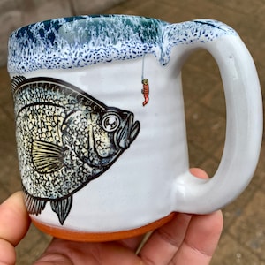 May include: White ceramic mug with a blue and white speckled glaze. The mug features a black and white illustration of a crappie fish with a red fishing lure hanging above it.