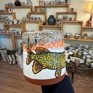 May include: A white ceramic mug with a brown rim and a hand-painted image of a yellow and green fish with black outlines. The mug has a speckled glaze with orange and green accents.