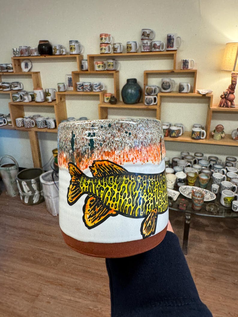May include: A white ceramic mug with a brown rim and a hand-painted image of a yellow and green fish with black outlines. The mug has a speckled glaze with orange and green accents.