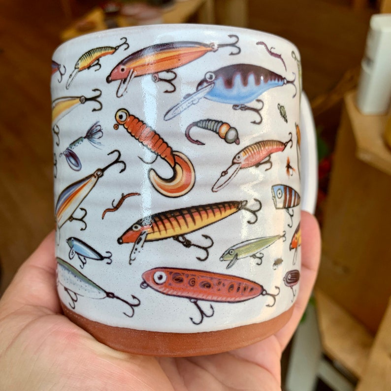 May include: White ceramic mug with a colorful pattern of fishing lures. The lures are in various colors and designs, including red, orange, yellow, blue, and green.