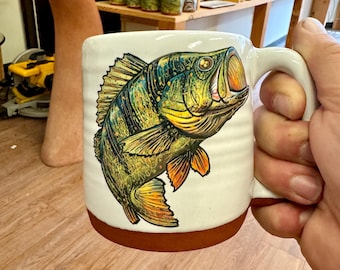 May include: A white ceramic mug with a brown rim features a colorful illustration of a largemouth bass with green, yellow, and orange details. The mug has a handle.