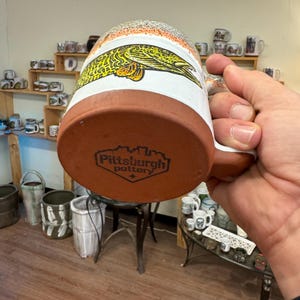 May include: A brown ceramic mug with a white glaze and a fish design. The mug has a brown handle and the bottom of the mug has the text "Pittsburgh Pottery" in black.