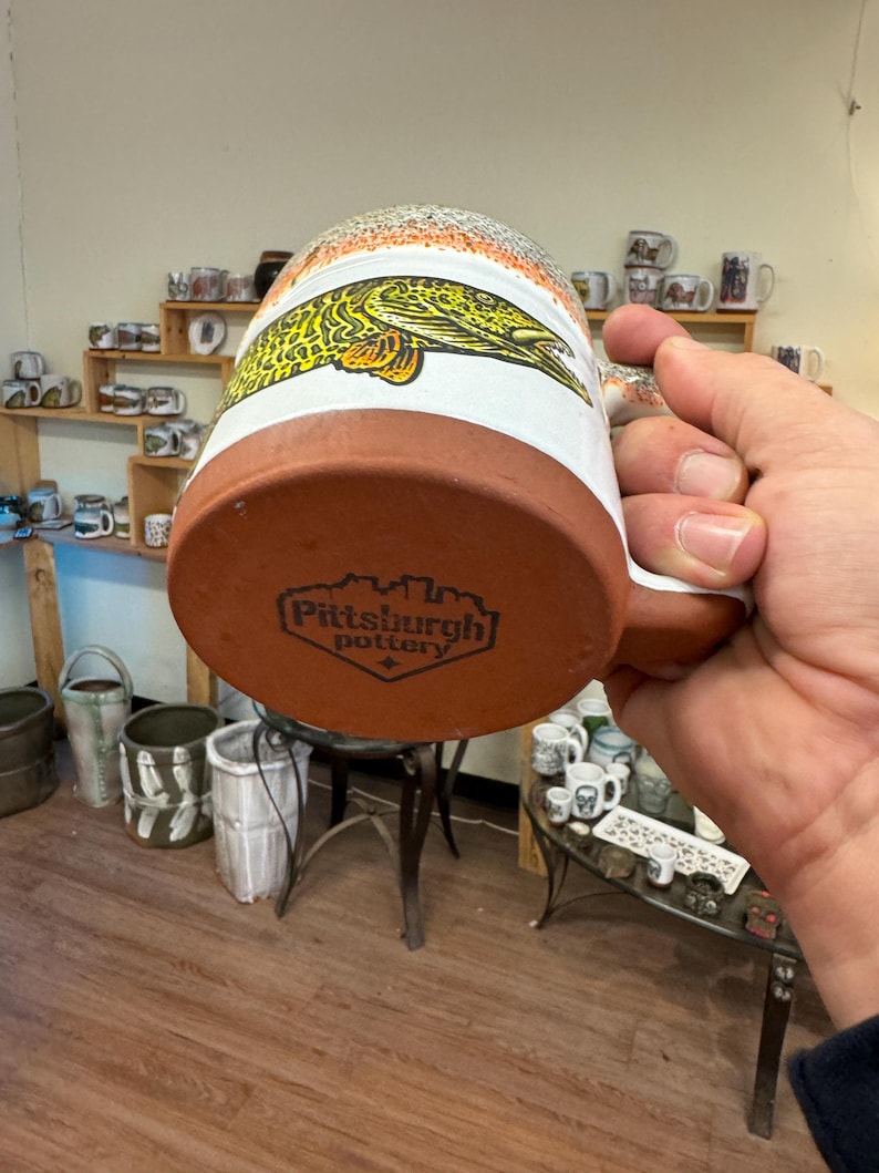 May include: A brown ceramic mug with a white glaze and a fish design. The mug has a brown handle and the bottom of the mug has the text "Pittsburgh Pottery" in black.