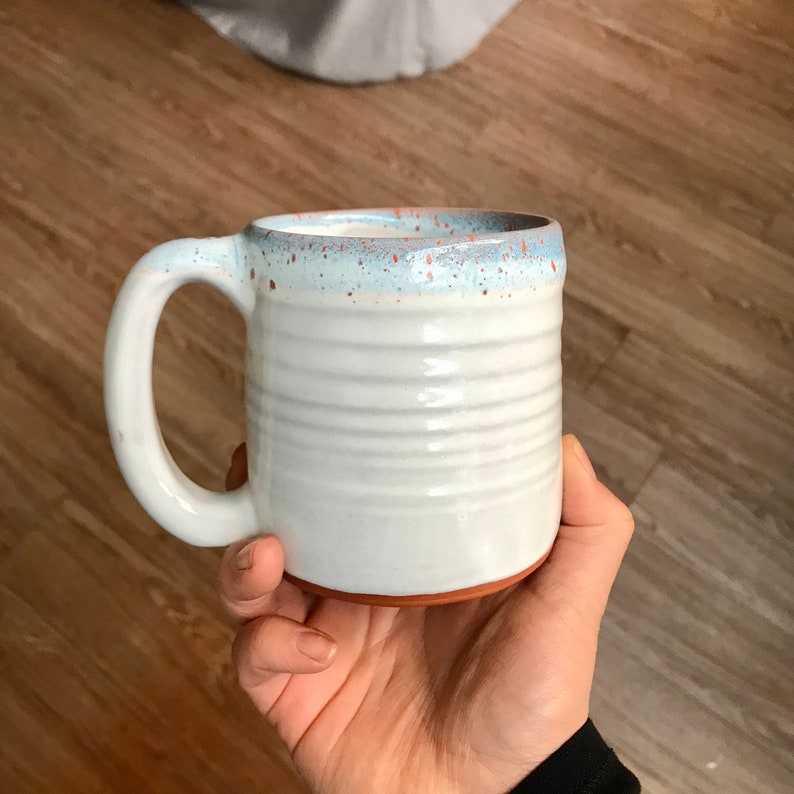 May include: White ceramic mug with a light blue and orange speckled rim. The mug has a rounded handle and a slightly flared lip.