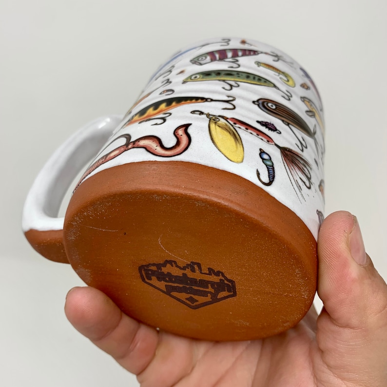 May include: White ceramic mug with a brown rim and a design of various fishing lures. The bottom of the mug has a brown stamp that reads "Pottery Barn".