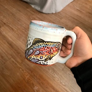 May include: White ceramic mug with a colorful trout illustration. The trout is swimming in a stream of water with blue, yellow, and orange accents. The mug has a handle and a light blue glaze.
