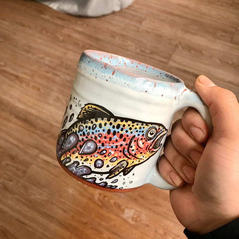 May include: A white ceramic mug with a brown, yellow, and orange trout design. The mug has a blue glaze and a speckled finish.