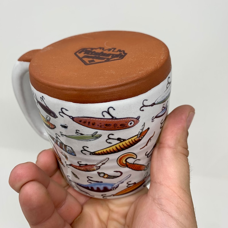 May include: A white ceramic mug with a brown rim and a brown handle. The mug is decorated with a pattern of colorful fishing lures. The mug has a brown logo on the top that says "Fishin' Friends".