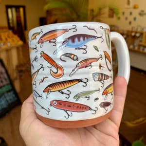 May include: A white ceramic mug with a brown rim features a colorful pattern of fishing lures. The lures are various colors and shapes, including red, orange, yellow, blue, and green.