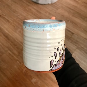 May include: White ceramic mug with a blue rim and a hand-painted design of purple, yellow, and brown swirls.