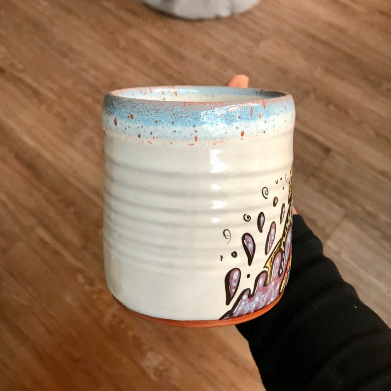 May include: White ceramic mug with a blue rim and a hand-painted design of purple, yellow, and brown swirls.