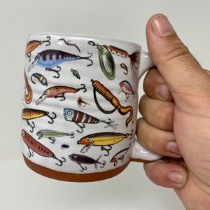 May include: A white ceramic mug with a brown rim, featuring a colorful pattern of fishing lures. The mug has a handle on the right side.