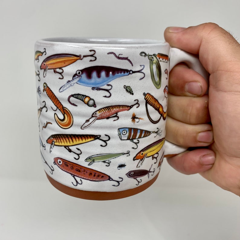 May include: A white ceramic mug with a brown rim features a colorful pattern of fishing lures. The lures are depicted in various colors and styles, including red, yellow, blue, and green.