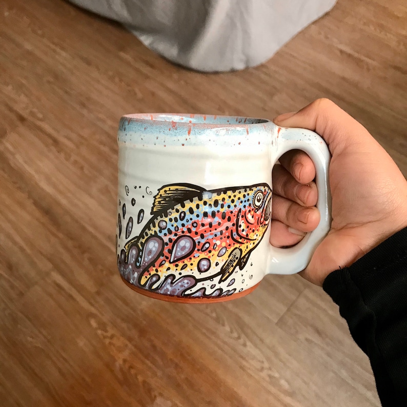 May include: A white ceramic mug with a handle features a colorful illustration of a trout swimming in water. The mug has a speckled glaze.