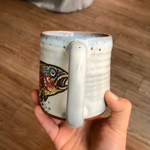 May include: White ceramic mug with a brown, red, yellow, and blue trout illustration. The mug has a speckled glaze and a brown rim.